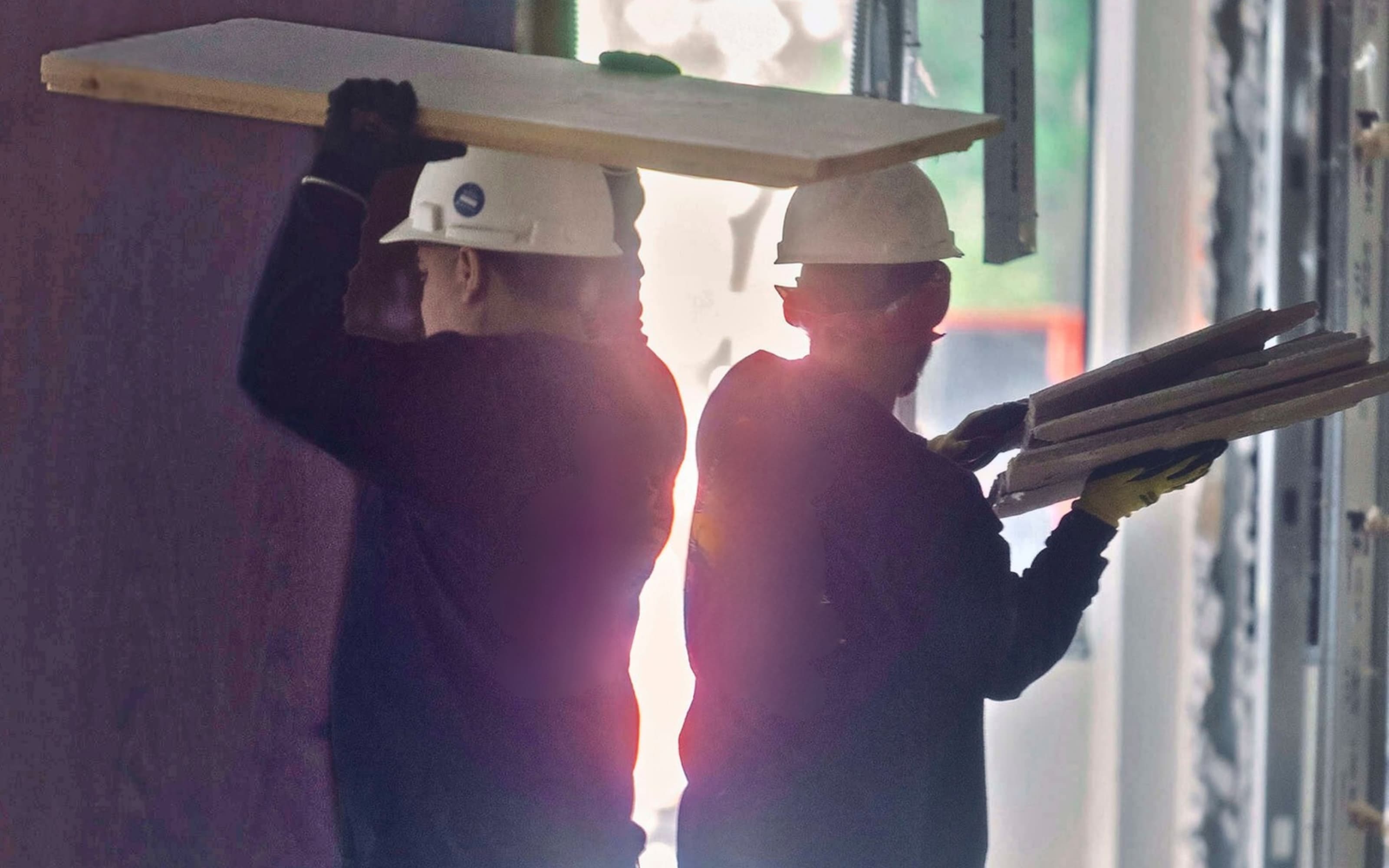 Two team members carrying panels on a backlit construction site